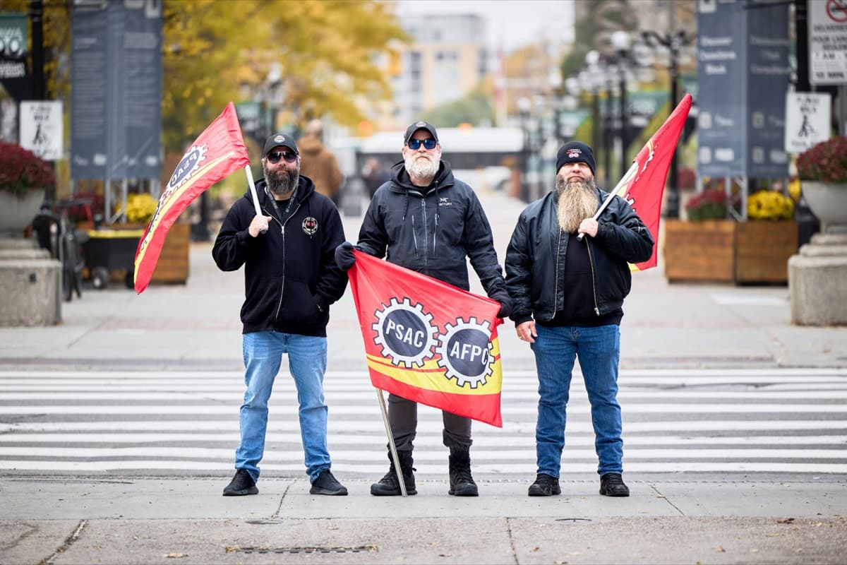 Three picketers stand on a crosswalk holding PSAC-AFPC flags.